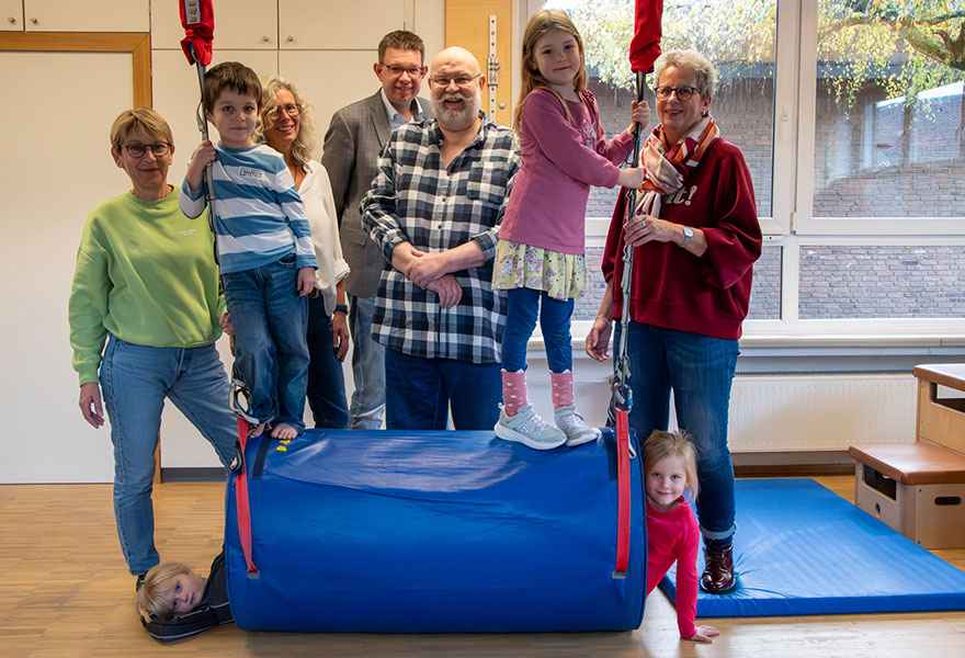 Erwachsene und Kinder stehen hinter bzw. auf einer großen blauen Rolle, die mit Seilen an der Decke befestigt ist. Erwachsene und Kinder stehen hinter bzw. auf einer großen blauen Rolle, die mit Seilen an der Decke befestigt ist.