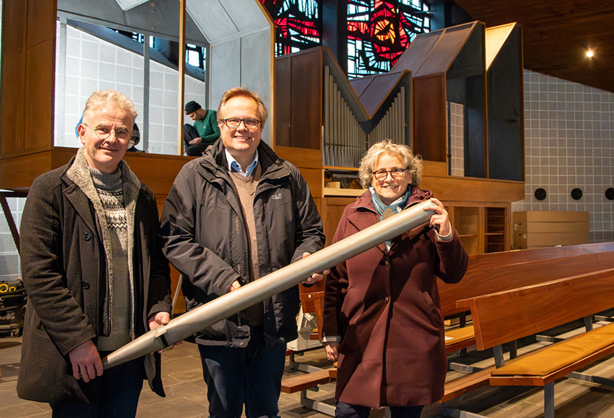 Kantor Thomas Drees, Pfarrer Michael Ostholthoff und Referentin Cäcilia Scholten halten eine große Orgelpfeife in der Hand. Hinter ihnen sieht man den Aufbau der neuen Orgel.