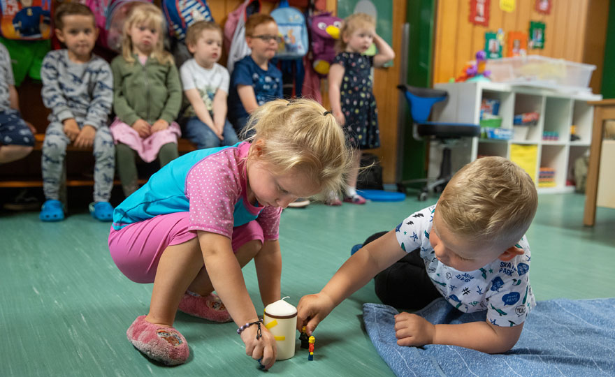 Zwei Kinder spielen in einem Kindergartenraum auf dem Boden mit einer Kerze und einer Figur. Im Hintergrund sind weitere Kinder zu sehen. 
