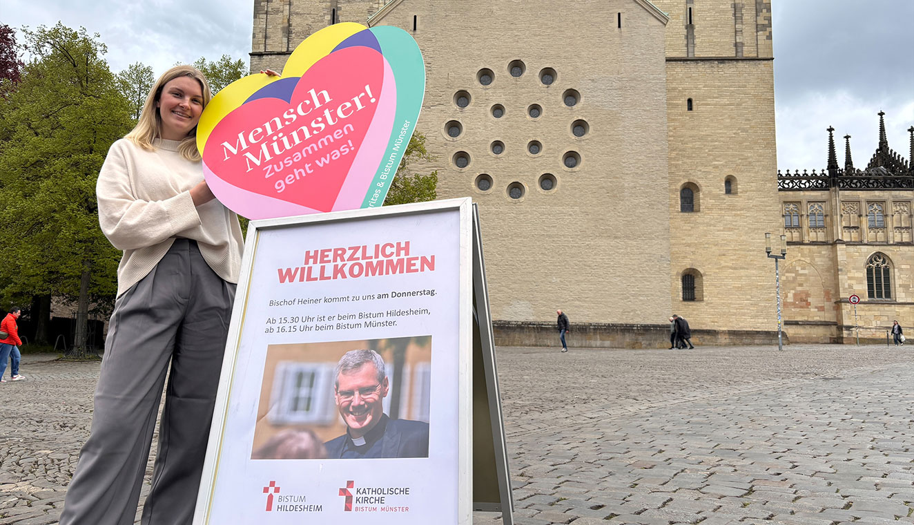 Marleen Michel steht vor dem St.-Paulus-Dom in Münster und hält ein buntes, herzförmiges Schild mit der Aufschrift „Mensch Münster! Zusammen geht was!“. Neben ihr steht ein Aufsteller mit der Überschrift „Herzlich willkommen“ und Informationen zu einem Besuch eines Bischofs.