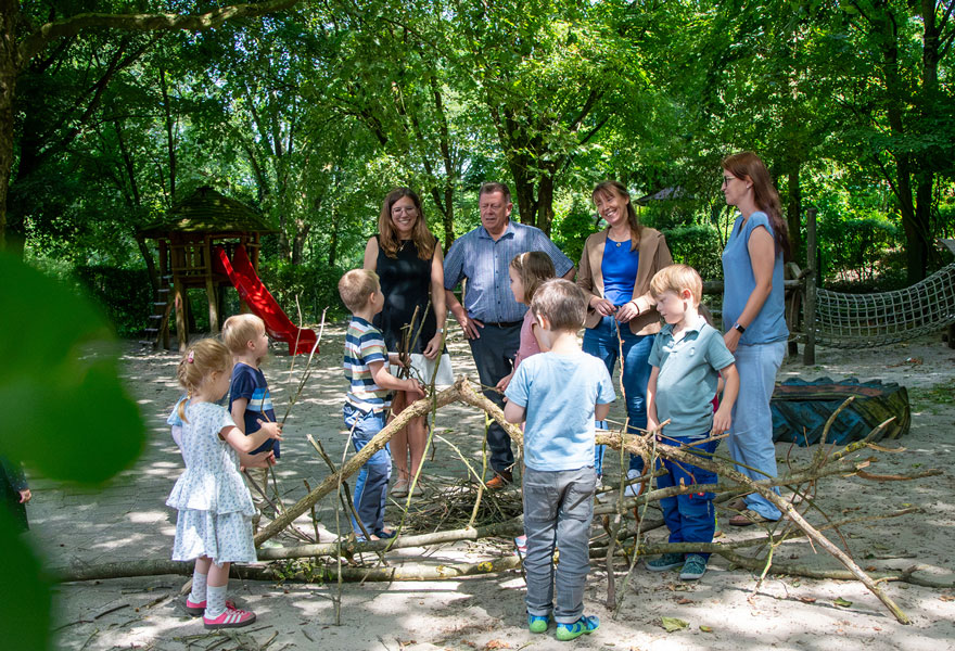 Im Vordergrund spielen Kinder mit Stöcken, dahinter stehen vier Erwachsene und schauen es sich an. Im Vordergrund spielen Kinder mit Stöcken, dahinter stehen vier Erwachsene und schauen es sich an.