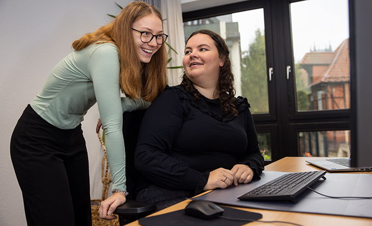 Eine junge Frau sitzt am Schreibtisch vor dem Computer und eine weitere steht neben ihr.