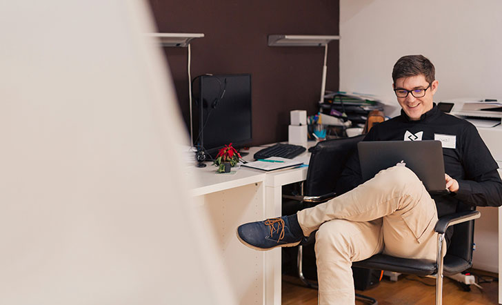 Ein junger Mann sitzt mit einem Laptop auf den Knien auf einem Bürostuhl.