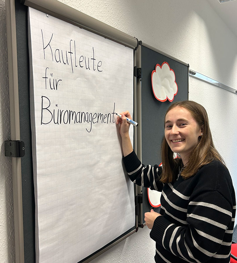 Sophie Messing steht mit einem Permanentmarker an einem Flipchart und schreibt: "Kaufleute für Büromanagement"