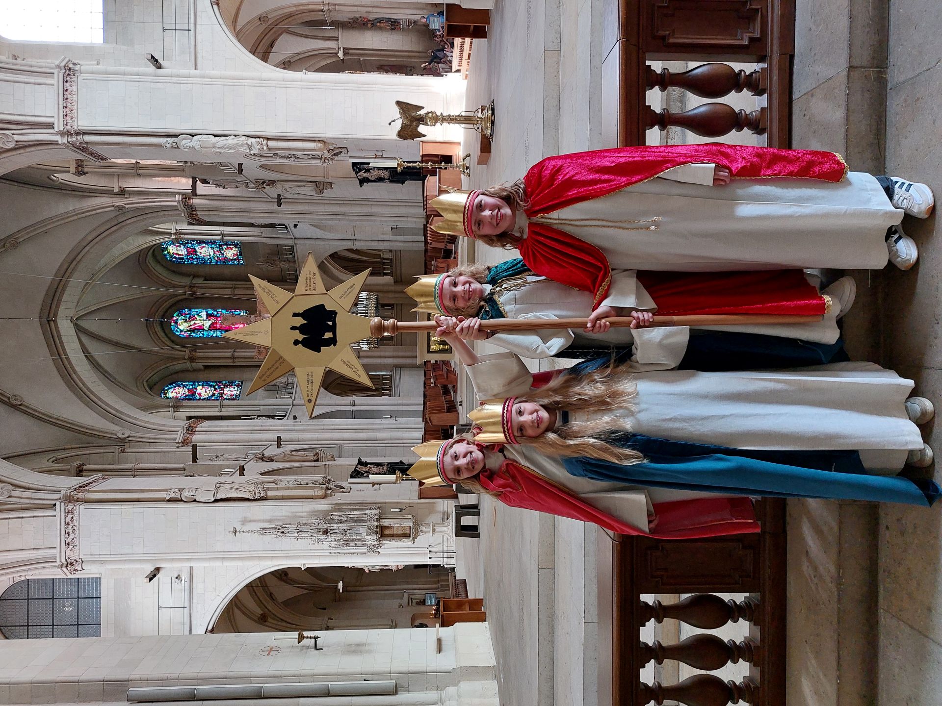 Vier als Sternsingerinnen verkleidete Kinder stehen lächelnd im St.-Paulus-Dom. Sie tragen weiße Gewänder, farbige Umhänge und goldene Kronen; eines der Kinder hält einen Stab mit einem großen goldenen Stern. Vier als Sternsingerinnen verkleidete Kinder stehen lächelnd im St.-Paulus-Dom. Sie tragen weiße Gewänder, farbige Umhänge und goldene Kronen; eines der Kinder hält einen Stab mit einem großen goldenen Stern.