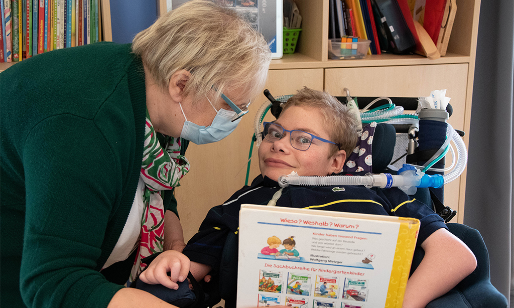Birgitt Gottfried zeigt ihrem Sohn Paul ein Buch. Paul sitzt im Rollstuhl, der Schlauch eines Beatmungsgerätes  ist an seinem Körper angeschlossen.