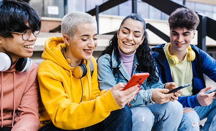 Vier junge Menschen sitzen nebeneinander und schauend lachend auf einen Handy-Bildschirm. 